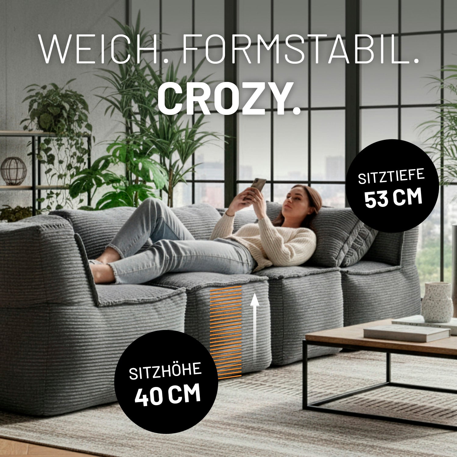 Modulare graue Lounge-Sitzlandschaft mit 40 cm Sitzhöhe und 53 cm Sitztiefe, auf der eine Frau entspannt liegend ihr Smartphone nutzt.