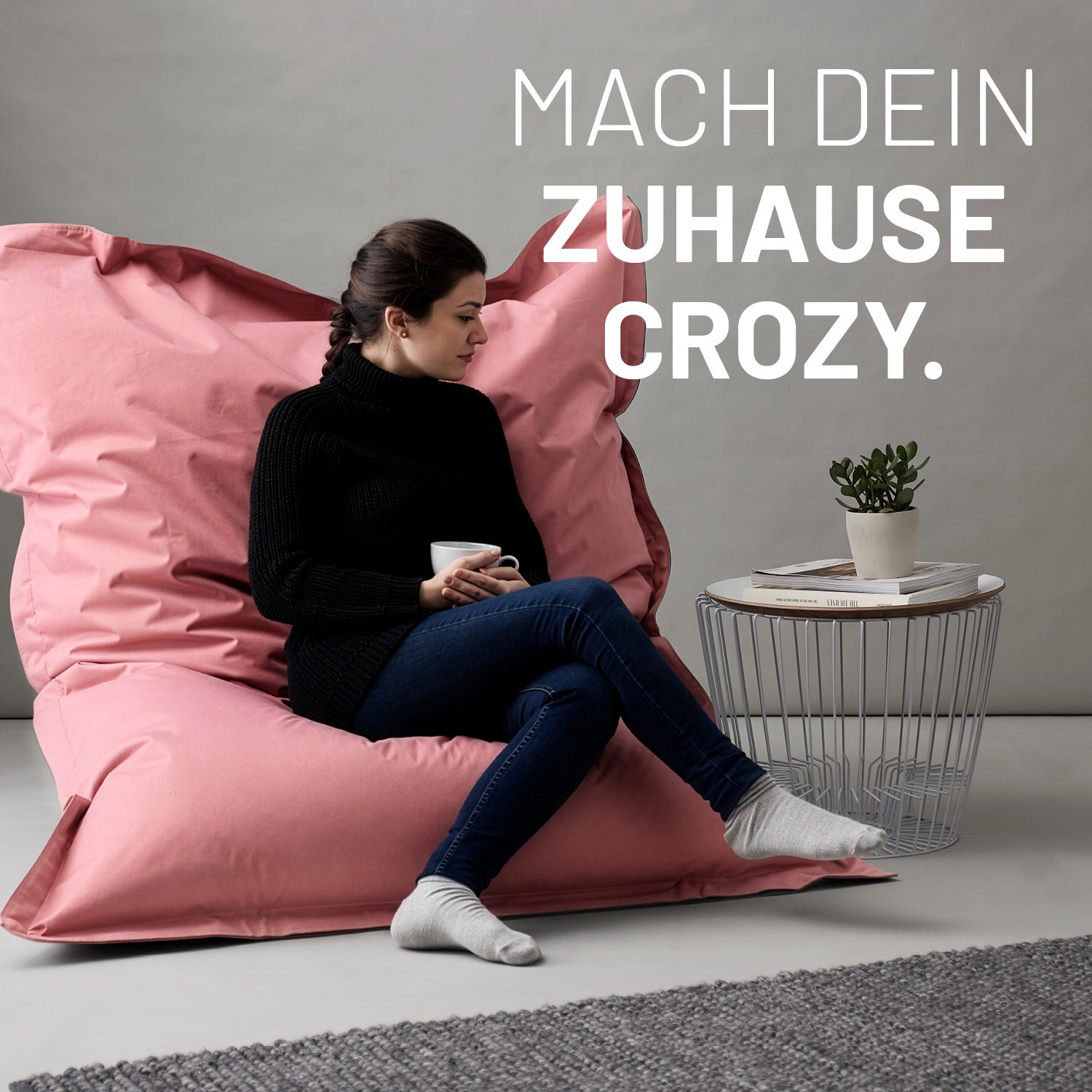 Eine Frau sitzt auf dem rosanen XXL Sitzsack, eine Tasse in der Hand, neben ihr ein Tisch - Mach dein Zuhause crozy.