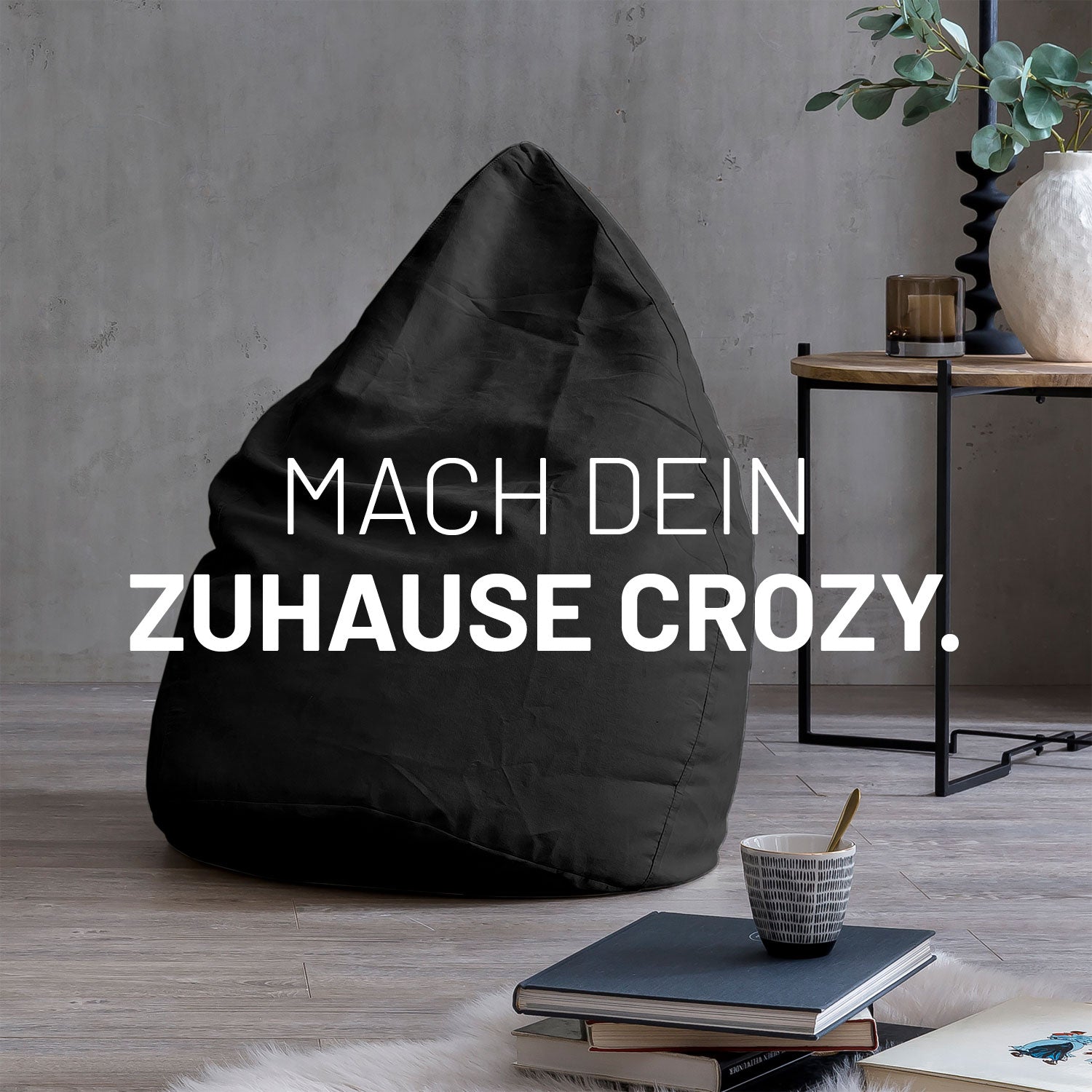 Großer Sitzsack in Schwarz in modernem Wohnambiente mit Tisch, Büchern und Tasse. Text: "Mach dein Zuhause crozy."
