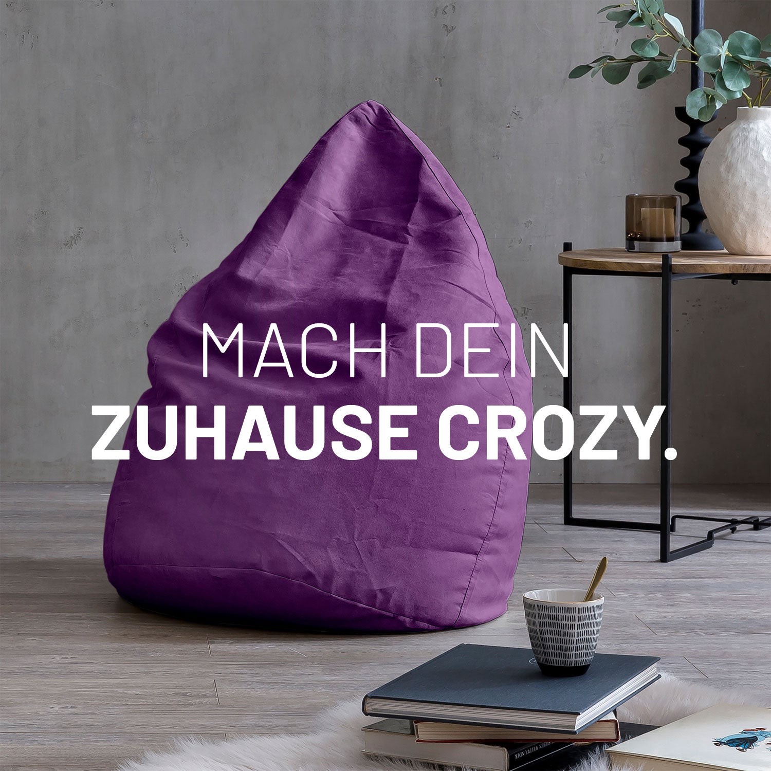 Großer Sitzsack in Lila in modernem Wohnambiente mit Tisch, Büchern und Tasse. Text: "Mach dein Zuhause crozy."