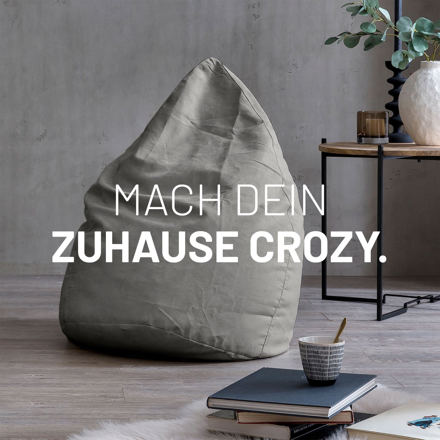 Großer Sitzsack in Grau in modernem Wohnambiente mit Tisch, Büchern und Tasse. Text: "Mach dein Zuhause crozy."