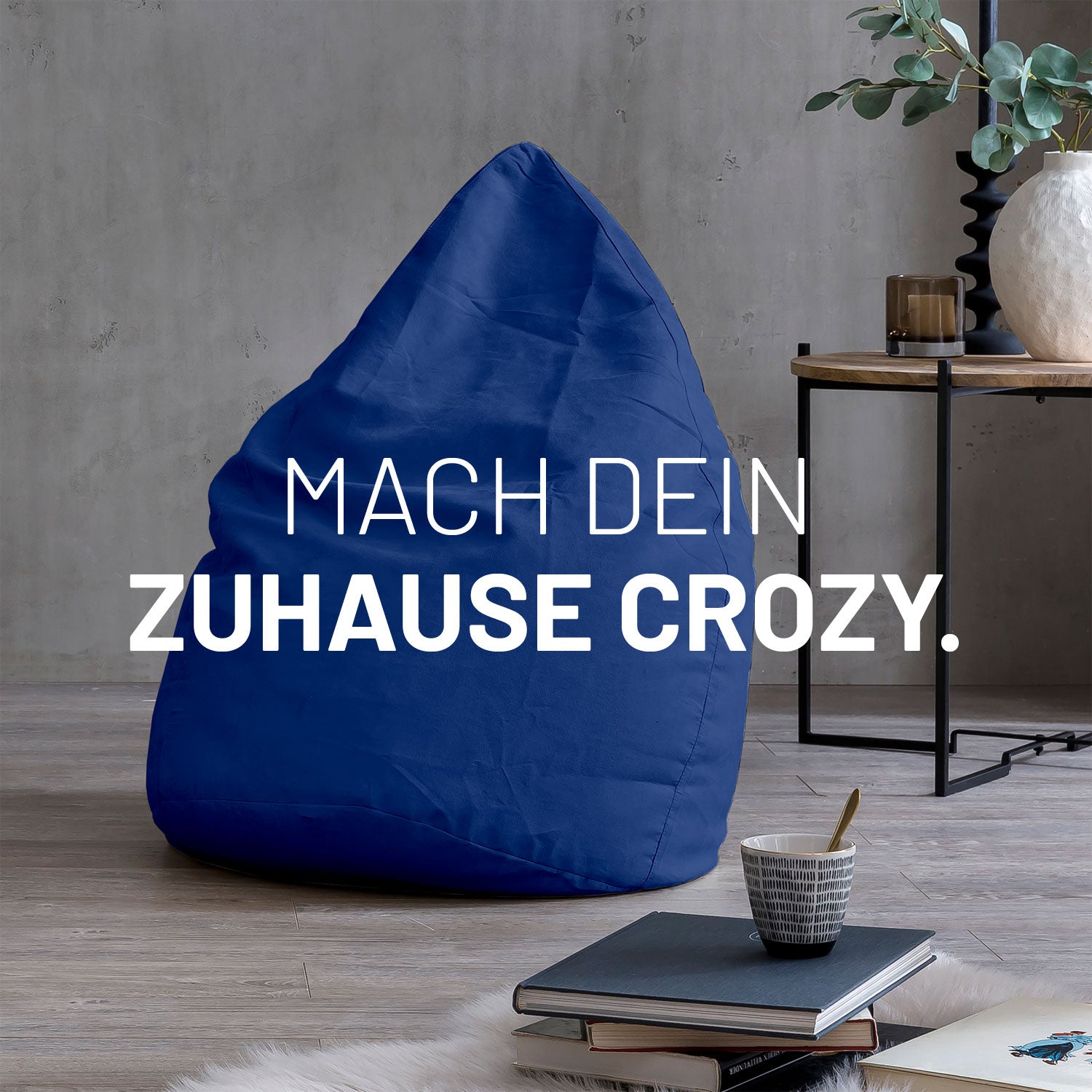 Großer Sitzsack in Royalblau in modernem Wohnambiente mit Tisch, Büchern und Tasse. Text: "Mach dein Zuhause crozy."
