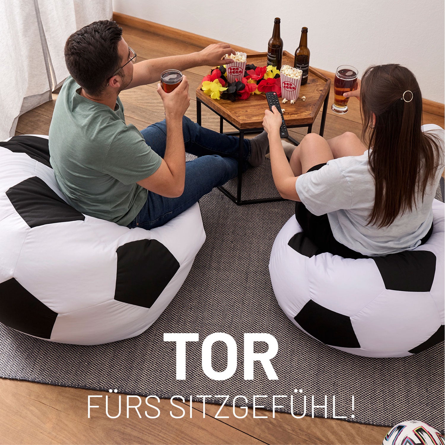 Fußball Fans sitzen auf den Fußball Sitzsäcken, vor ihnen ein Tisch mit Getränken, Snacks und Deutschland Hawaii Kette.