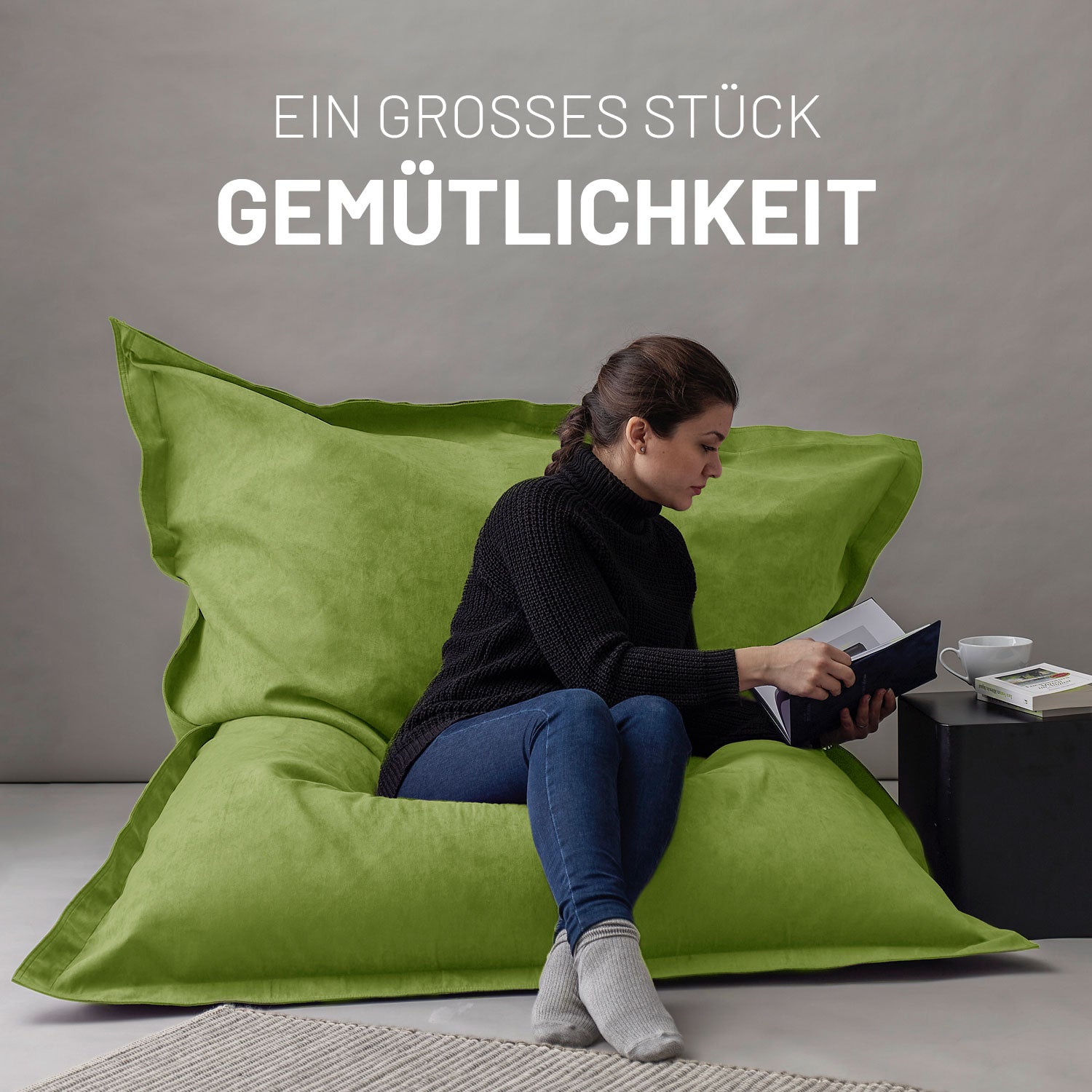Frau sitzt entspannt auf dem XXL Sitzsack Microvelour in Grün, liest ein Buch, daneben ein Tisch mit Tasse und Büchern, Schriftzug „Ein großes Stück Gemütlichkeit“.