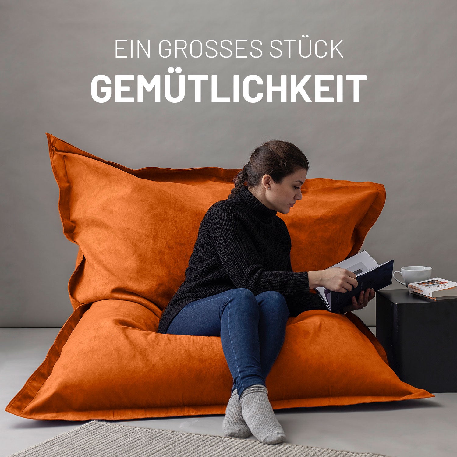 Frau sitzt entspannt auf dem XXL Sitzsack Microvelour in Orange, liest ein Buch, daneben ein Tisch mit Tasse und Büchern, Schriftzug „Ein großes Stück Gemütlichkeit“.