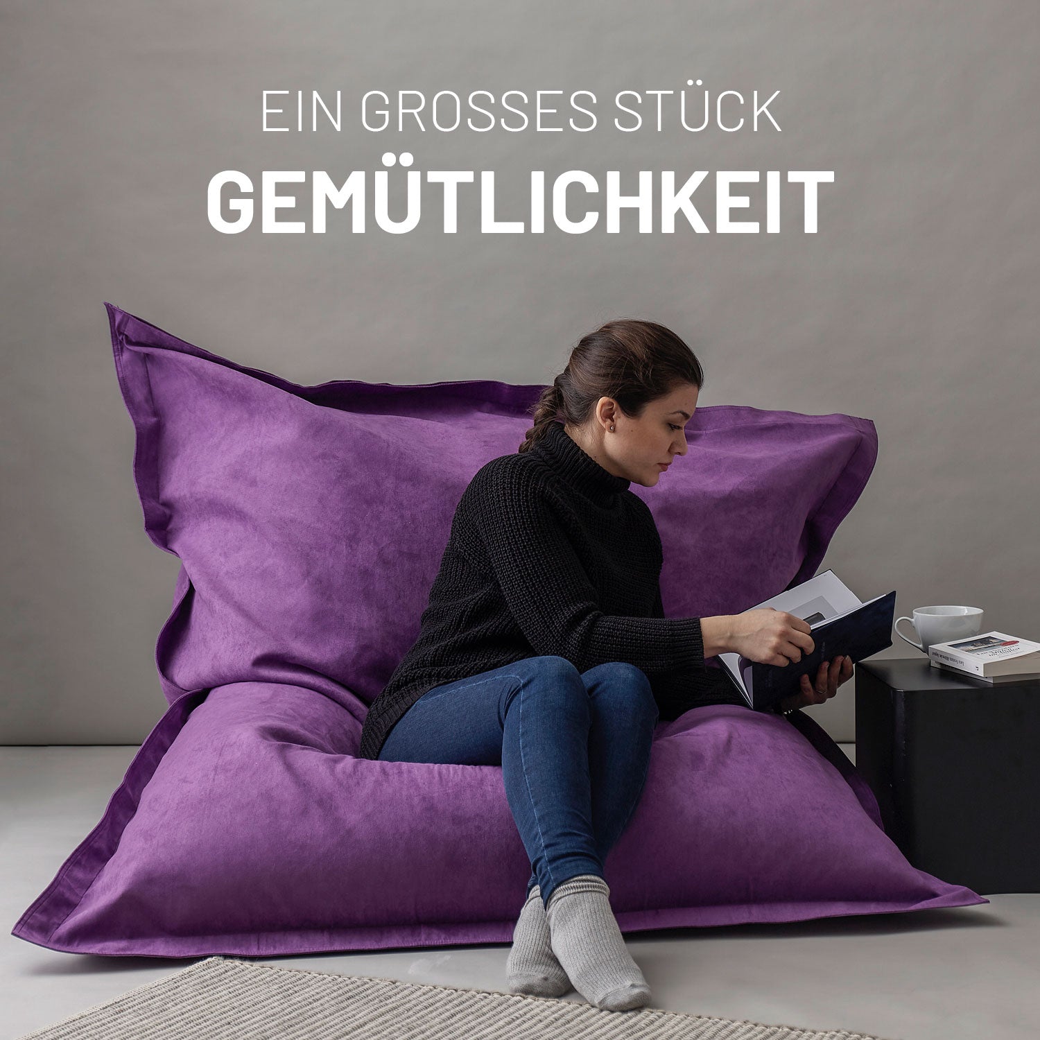 Frau sitzt entspannt auf dem XXL Sitzsack Microvelour in Lila, liest ein Buch, daneben ein Tisch mit Tasse und Büchern, Schriftzug „Ein großes Stück Gemütlichkeit“.