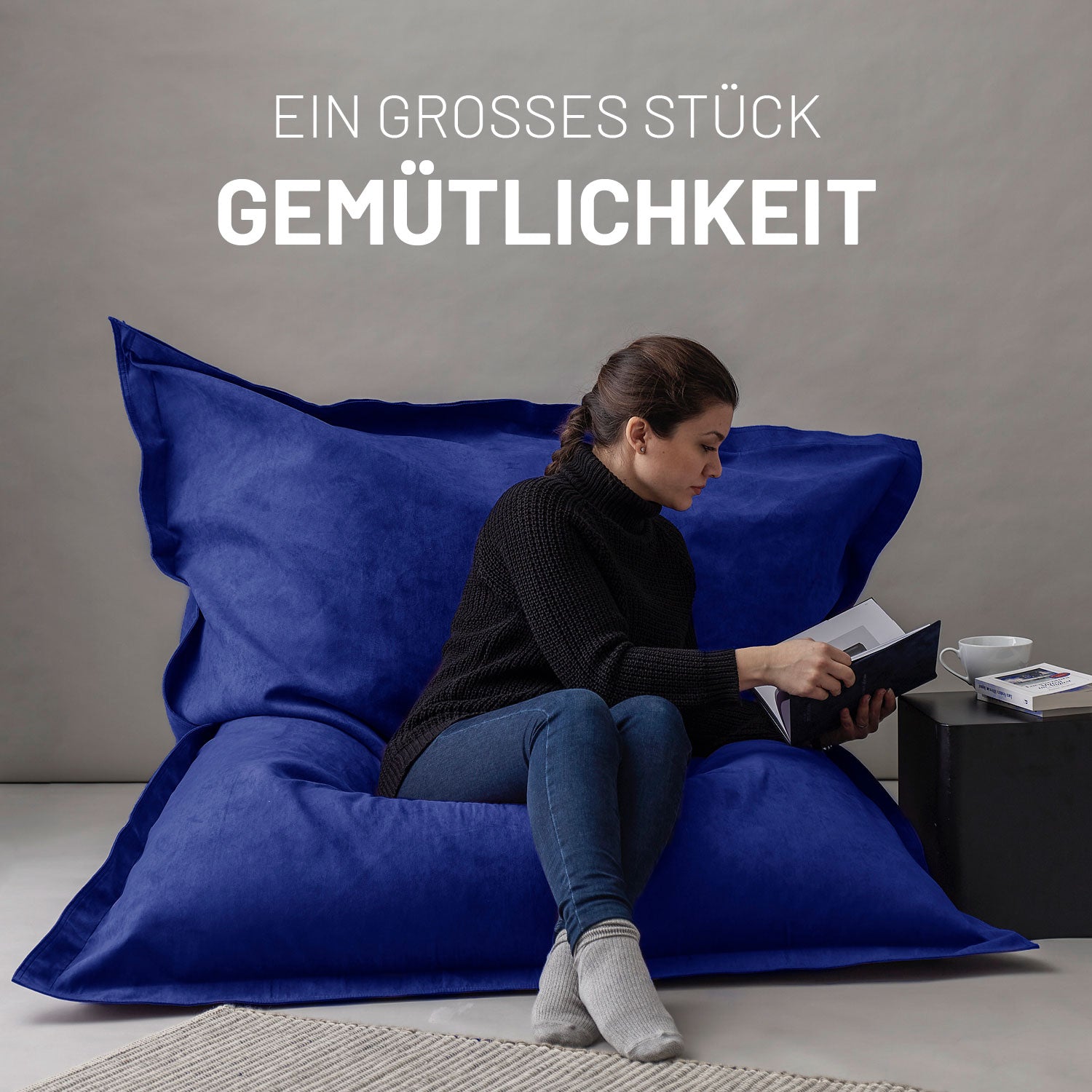 Frau sitzt entspannt auf dem XXL Sitzsack Microvelour in Royalblau, liest ein Buch, daneben ein Tisch mit Tasse und Büchern, Schriftzug „Ein großes Stück Gemütlichkeit“.