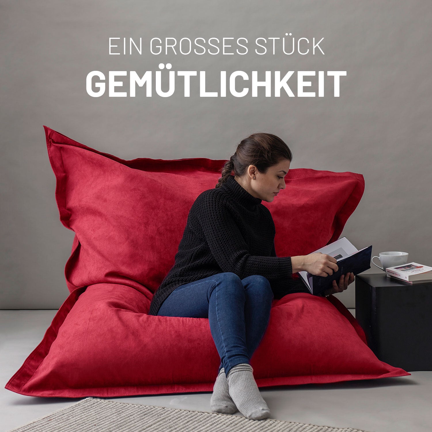 Frau sitzt entspannt auf dem XXL Sitzsack Microvelour in Rot, liest ein Buch, daneben ein Tisch mit Tasse und Büchern, Schriftzug „Ein großes Stück Gemütlichkeit“.