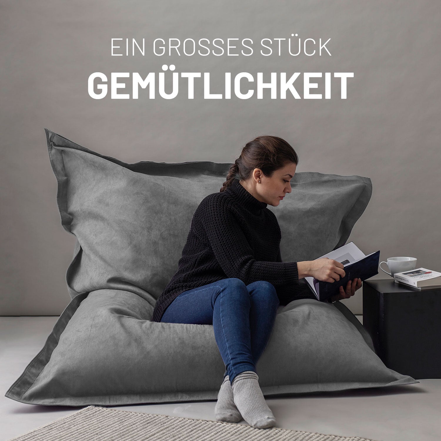 Frau sitzt entspannt auf dem XXL Sitzsack Microvelour in Grau, liest ein Buch, daneben ein Tisch mit Tasse und Büchern, Schriftzug „Ein großes Stück Gemütlichkeit“.