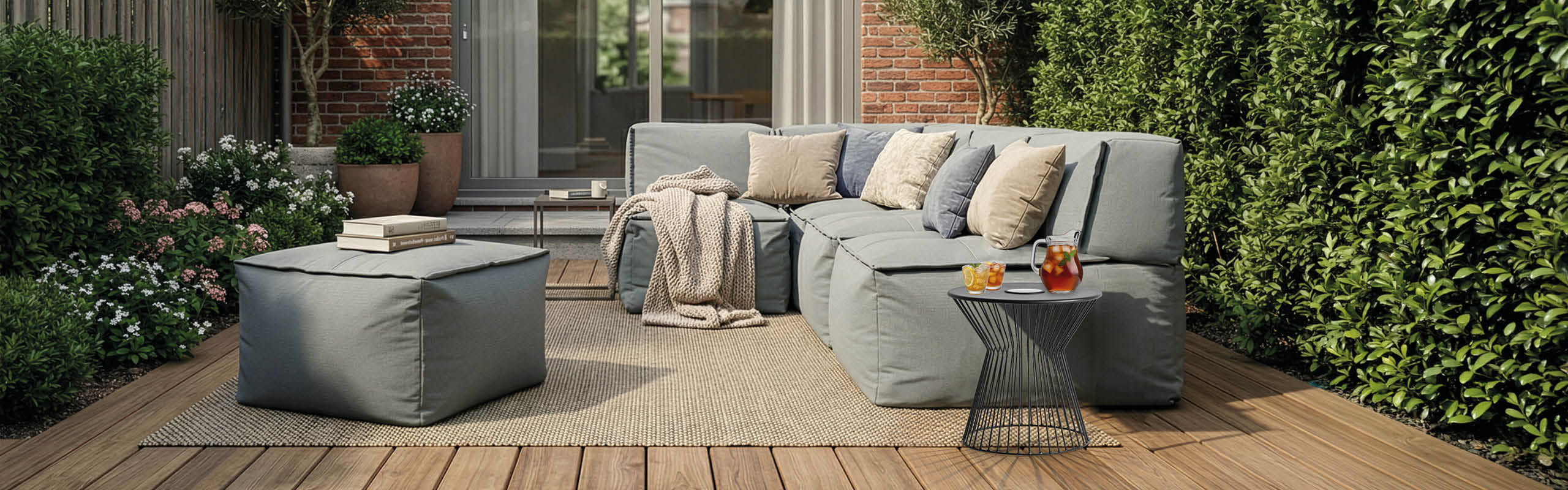 Modernes Outdoor-Wohnzimmer mit modularem Loungesofa und Hocker auf der Terrasse, ergänzt durch Kissen, Decke und sommerliche Deko.
