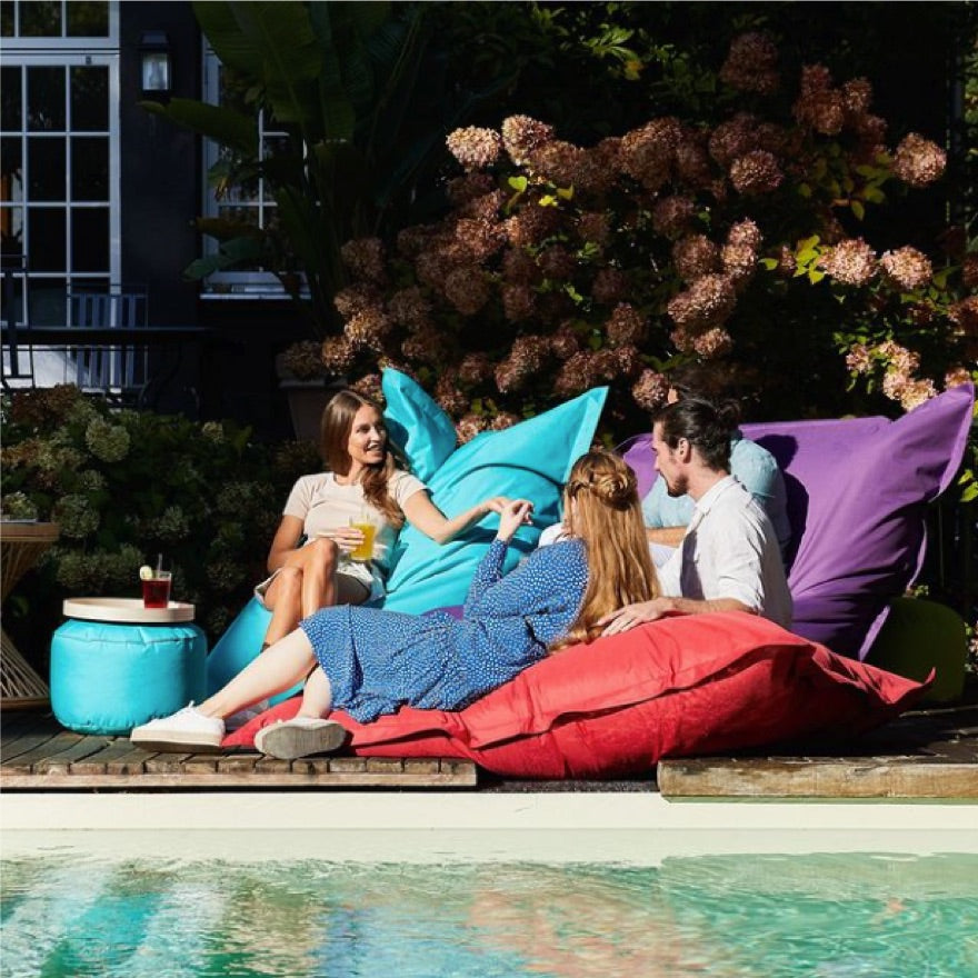 Frauen und Männer liegen im Sommer an einem Pool auf verschiedenen XXL Sitzsäcken, daneben ein Pouf als Ablage.
