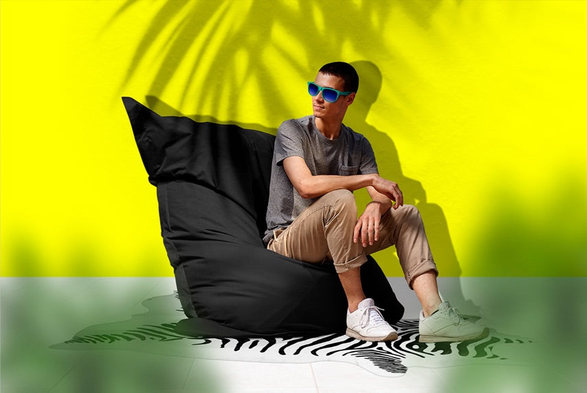 Ein Mann mit Sonnenbrille sitzt in sommerlicher Atmosphäre vor gelber Wand mit Palmenschatten auf dem schwarzen XXL Sitzsack.