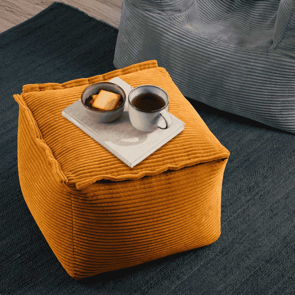 Gelber Cord-Hocker dient als Ablage für Tablett mit Tasse Kaffee und Keks, steht auf dunklem Teppich neben grauem Sitzsack in gemütlicher Wohnumgebung.