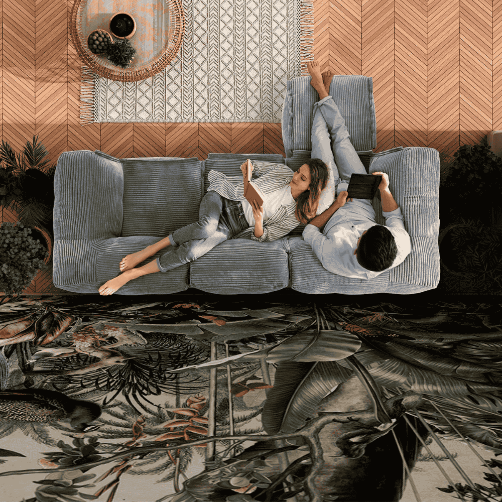 Paar liegt entspannt auf einem grauen Cord-Sofa, sie liest ein Buch, er hält ein Tablet; wohnliche Szene mit gemustertem Teppich und Holzboden.