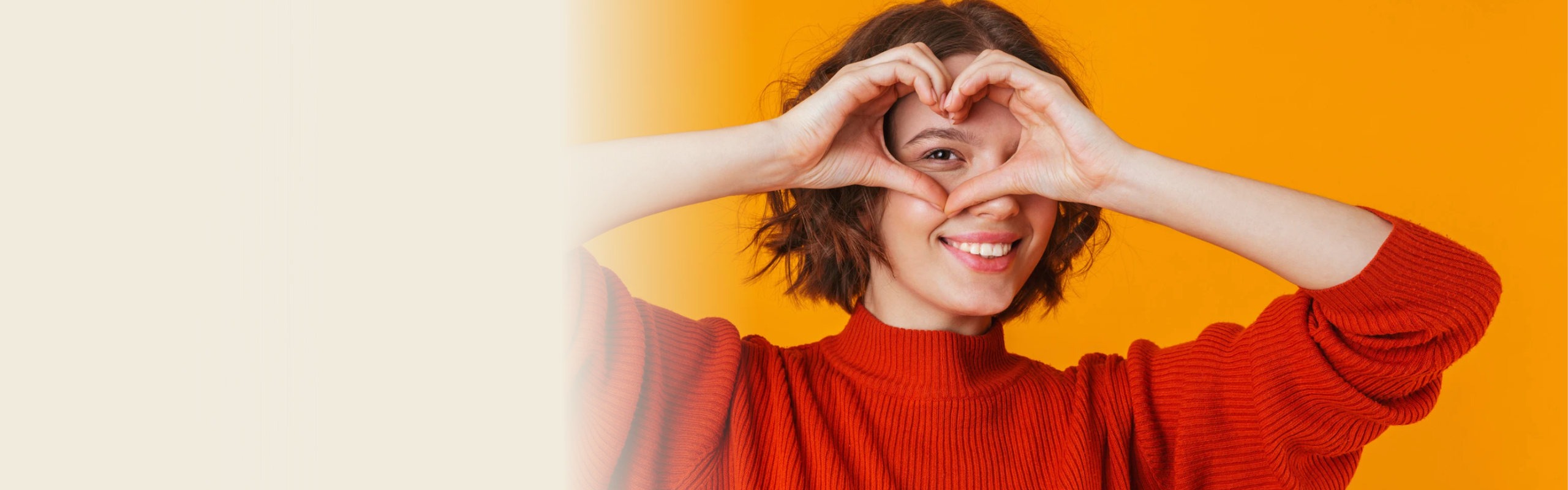 Frau mit kurzem braunen Haar und rotem Pullover formt ein Herz mit den Händen vor einem orangefarbenen Hintergrund und lächelt fröhlich.