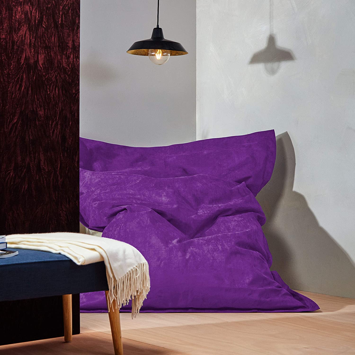 Der XXL Microvelours Sitzsack in Lila liegt in der Ecke eines Wohnzimmer, oben drüber eine Lampe, davor ein Tisch.