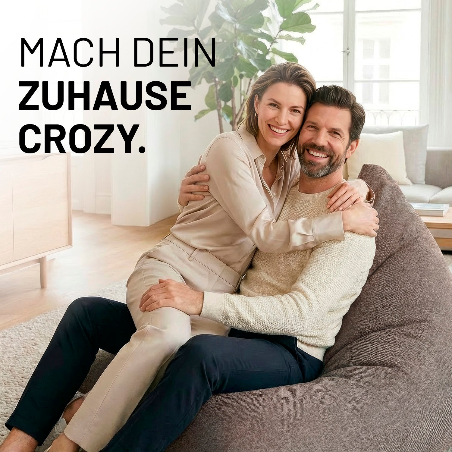 Ein lachendes Paar umarmt sich auf dem Interior Line Sitzsack in Hellgrau im hellen Wohnzimmer; Schriftzug „Mach dein Zuhause crozy.“