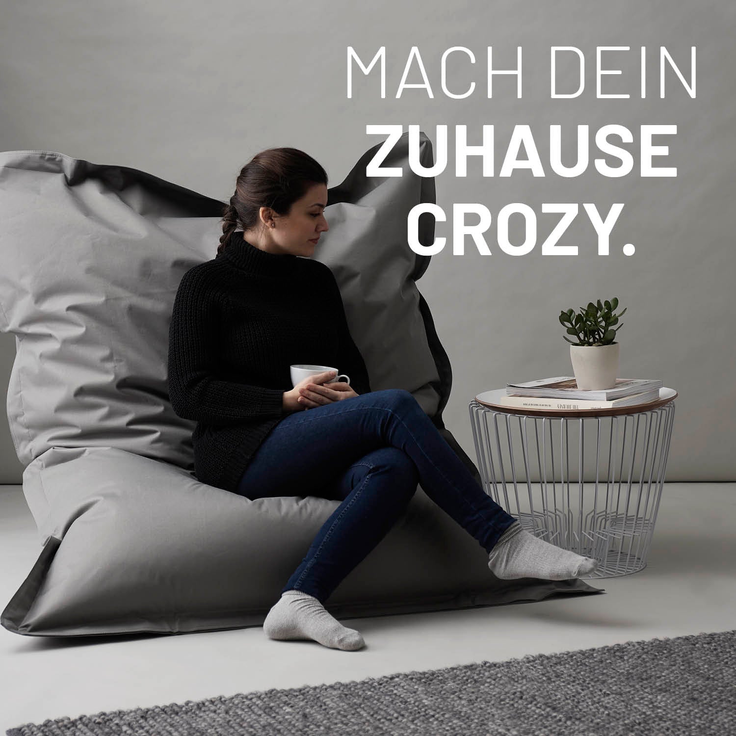 Eine Frau sitzt auf dem grauen XXL Sitzsack, eine Tasse in der Hand, neben ihr ein Tisch - Mach dein Zuhause crozy.