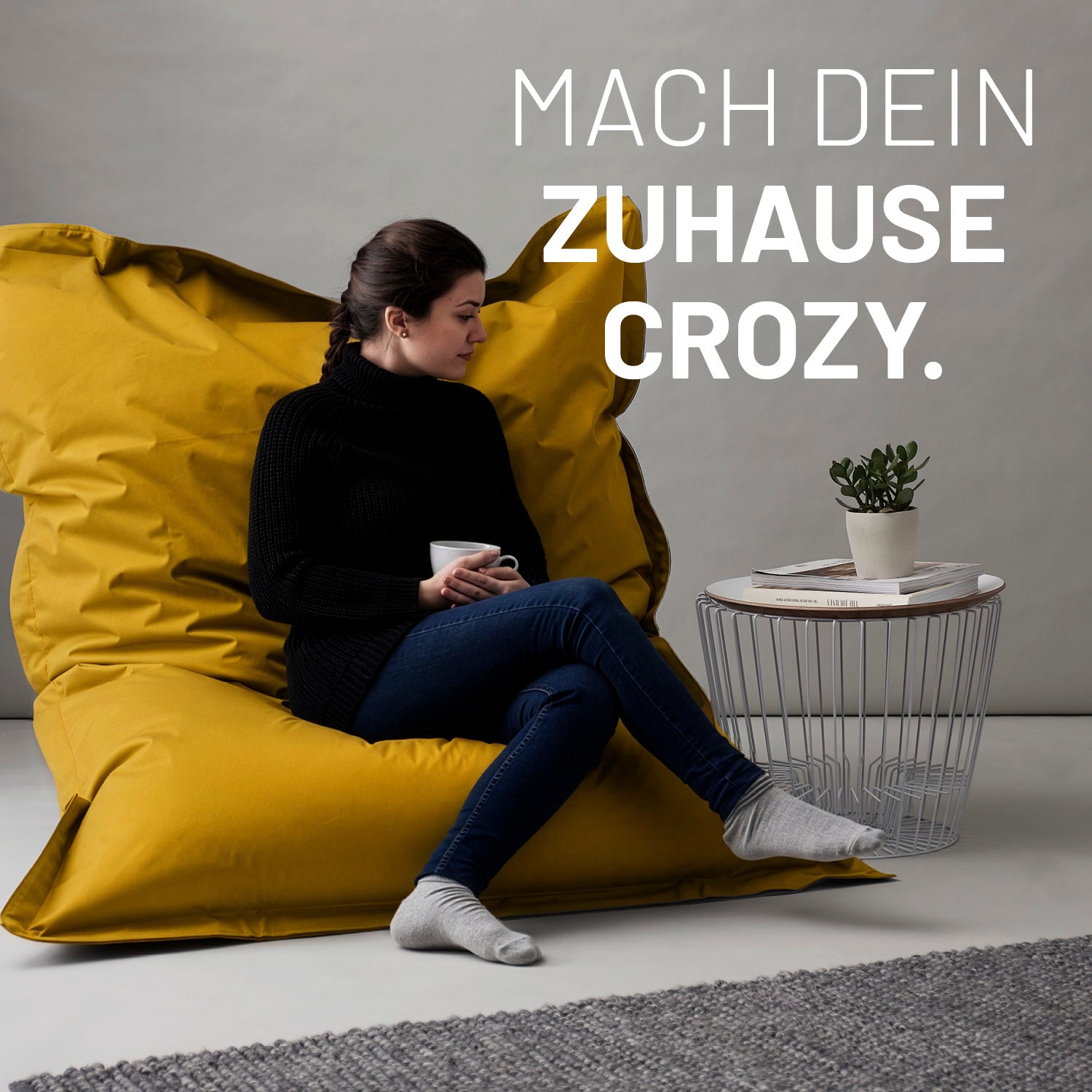 Eine Frau sitzt auf dem senfgelben XXL Sitzsack, eine Tasse in der Hand, neben ihr ein Tisch - Mach dein Zuhause crozy.