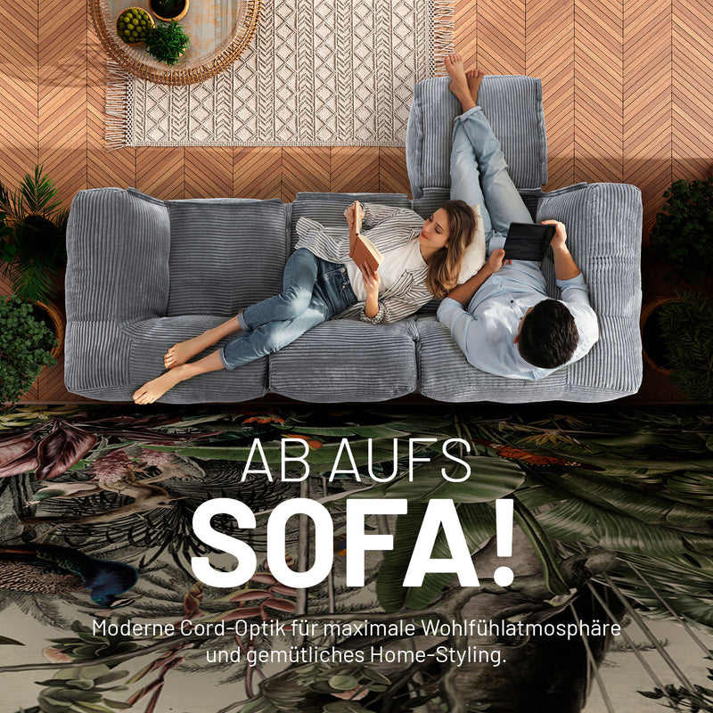 Frau mit Buch und Mann mit Tablet enspannen auf dem grauen Cord-Sitzsack-Sofa in einem modernen Wohnzimmer aus der Vogelperspektive. 