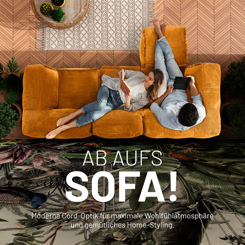 Frau mit Buch und Mann mit Tablet enspannen auf dem senfgelben Cord-Sitzsack-Sofa in einem modernen Wohnzimmer aus der Vogelperspektive. 