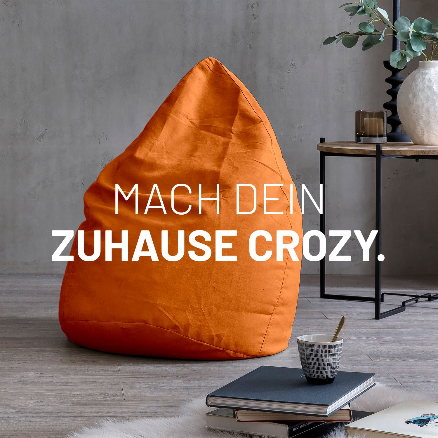XL Sitzsack in Orange in modernem Wohnambiente mit Tisch, Büchern und Tasse. Text: "Mach dein Zuhause crozy."