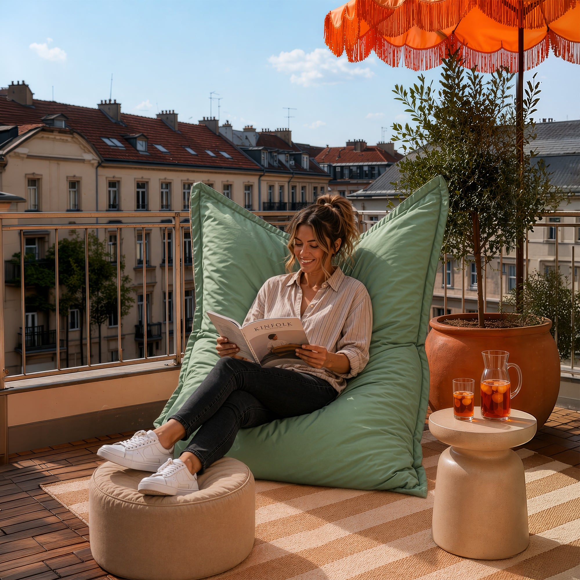 Frau sitzt auf pastellgrünem Sitzsack auf dem Balkon, liest entspannt ein Buch, Füße auf Pouf, sommerliche Szene mit Drinks und Sonnenschirm.
