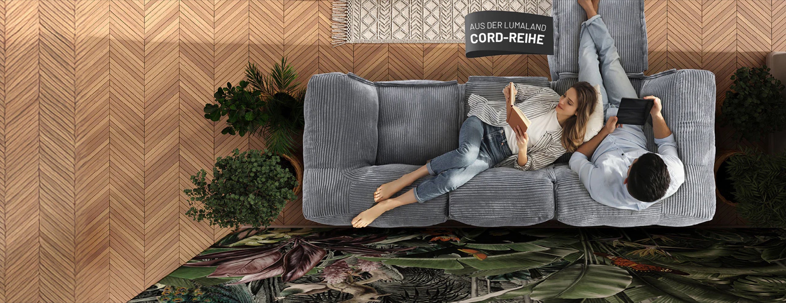 Frau mit Buch und Mann mit Tablet enspannen auf dem grauen Cord-Sitzsack-Sofa in einem modernen Wohnzimmer aus der Vogelperspektive. 