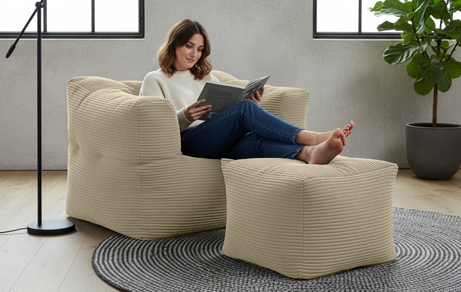 Frau liest entspannt auf beigem Cord-Sessel mit passendem Hocker in modernem Wohnzimmer, weich gepolstert und gemütlich.