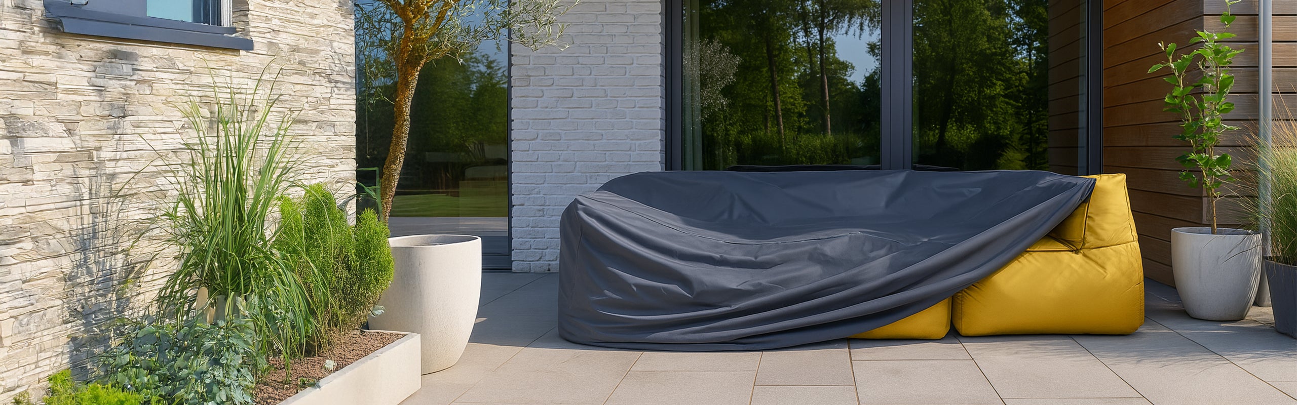 Gelbes LUMALAND Outdoor-Sofa auf Terrasse mit grauer Wetterschutzhaube, umgeben von Pflanzkübeln und moderner Hausfassade mit Stein- und Holzelementen.