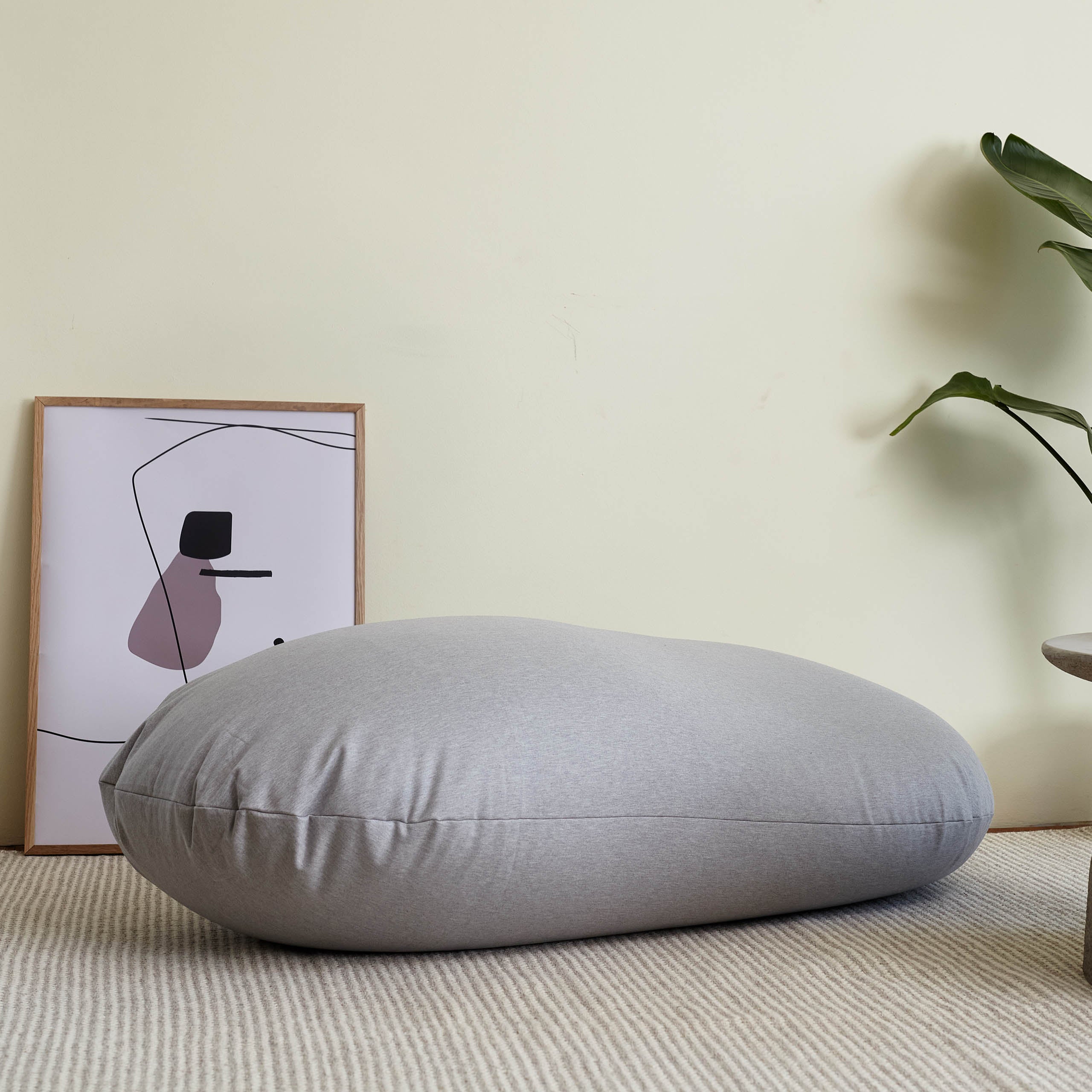 Flexi Comfort Sitzsack in Hellgrau als Liegekissen vor einer Wand in einem lichtdurchfluteten Wohnzimmer.