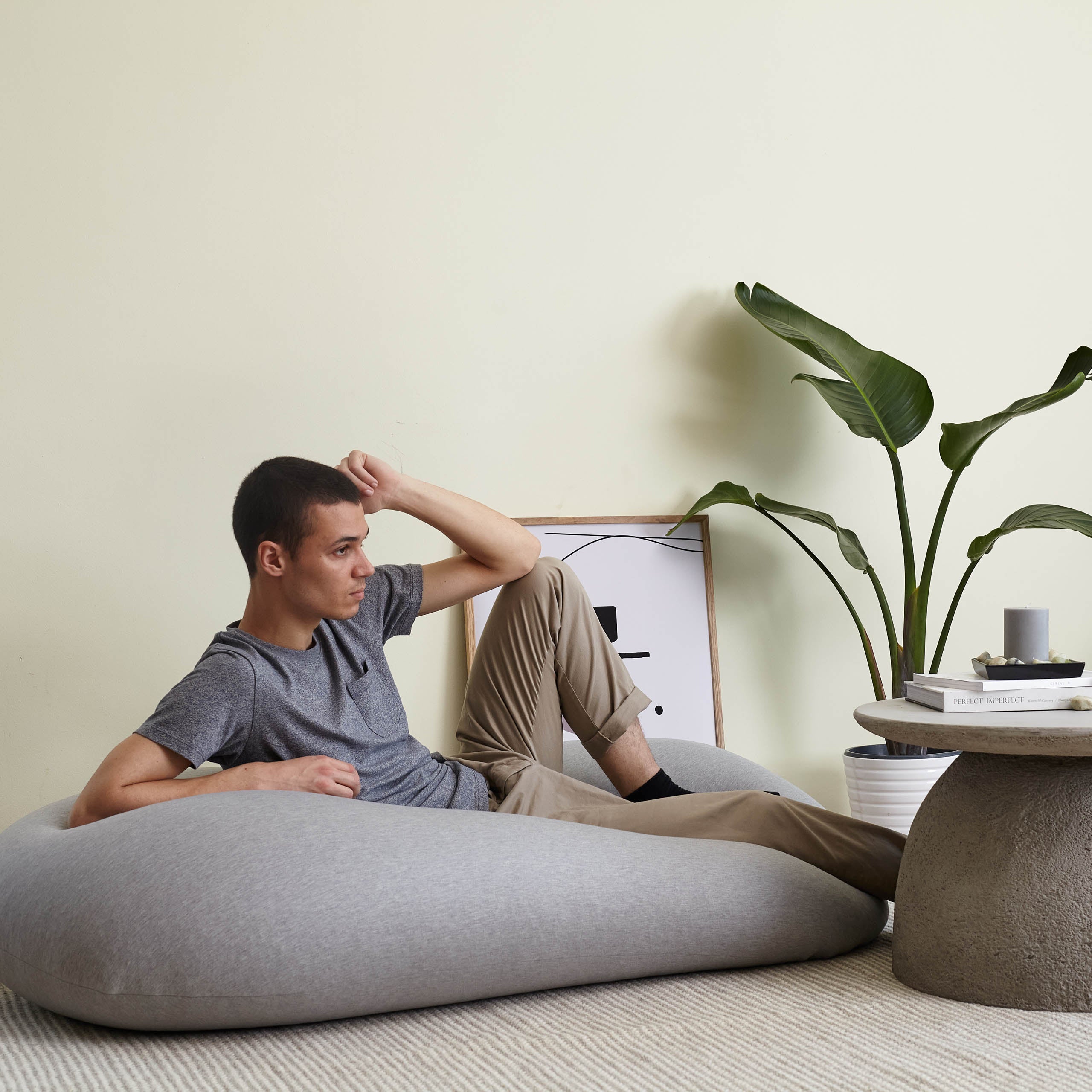 Ein Mann liegt mit aufgestelltem Bein auf dem hellgrauen Flexi Comfort Liege-Sitzsack, daneben ein Bild und ein Tisch.