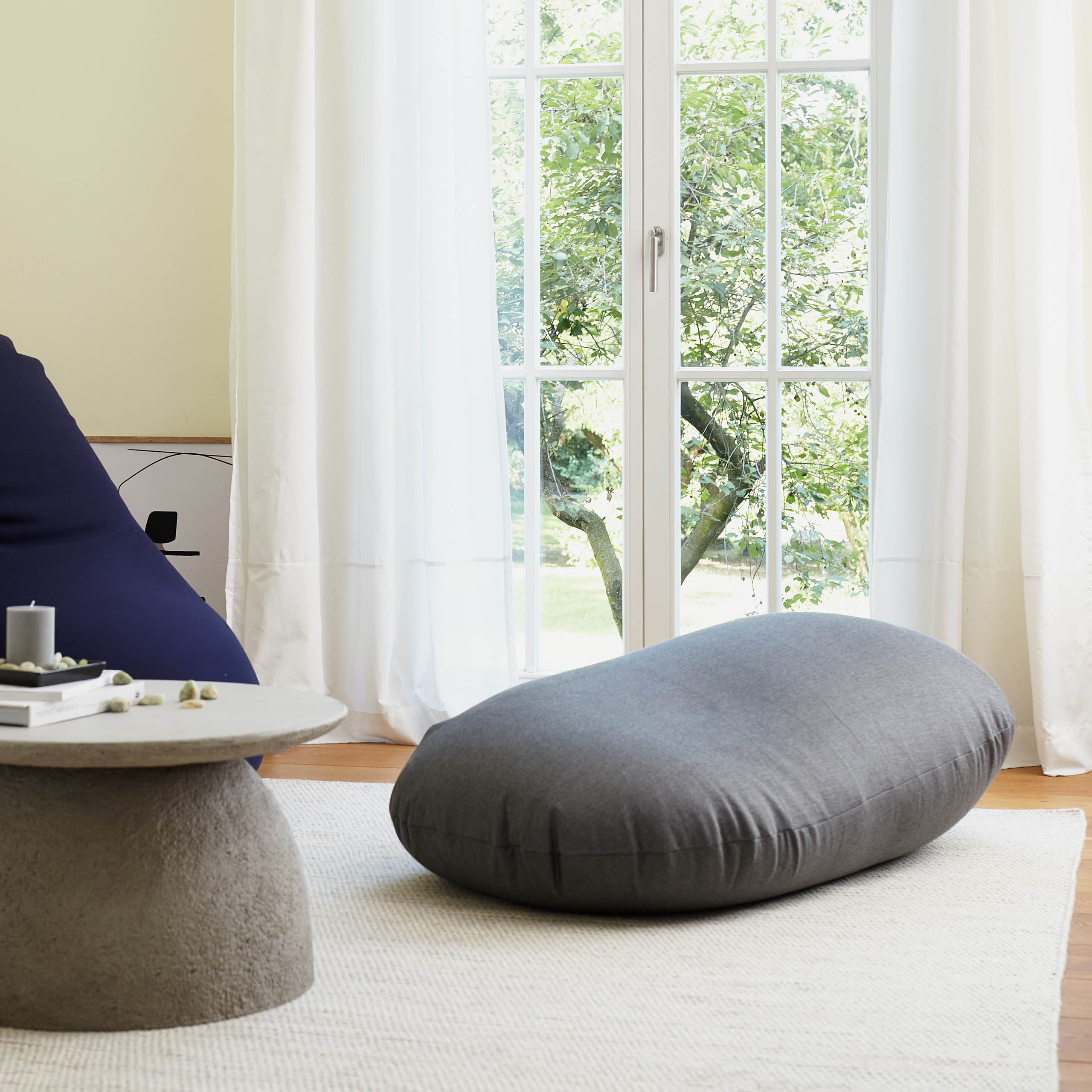 Flexi Comfort Sitzsack in Dunkelgrau als Liegekissen, am Rand der Sitzsack in Navyblau in einem lichtdurchfluteten Wohnzimmer.