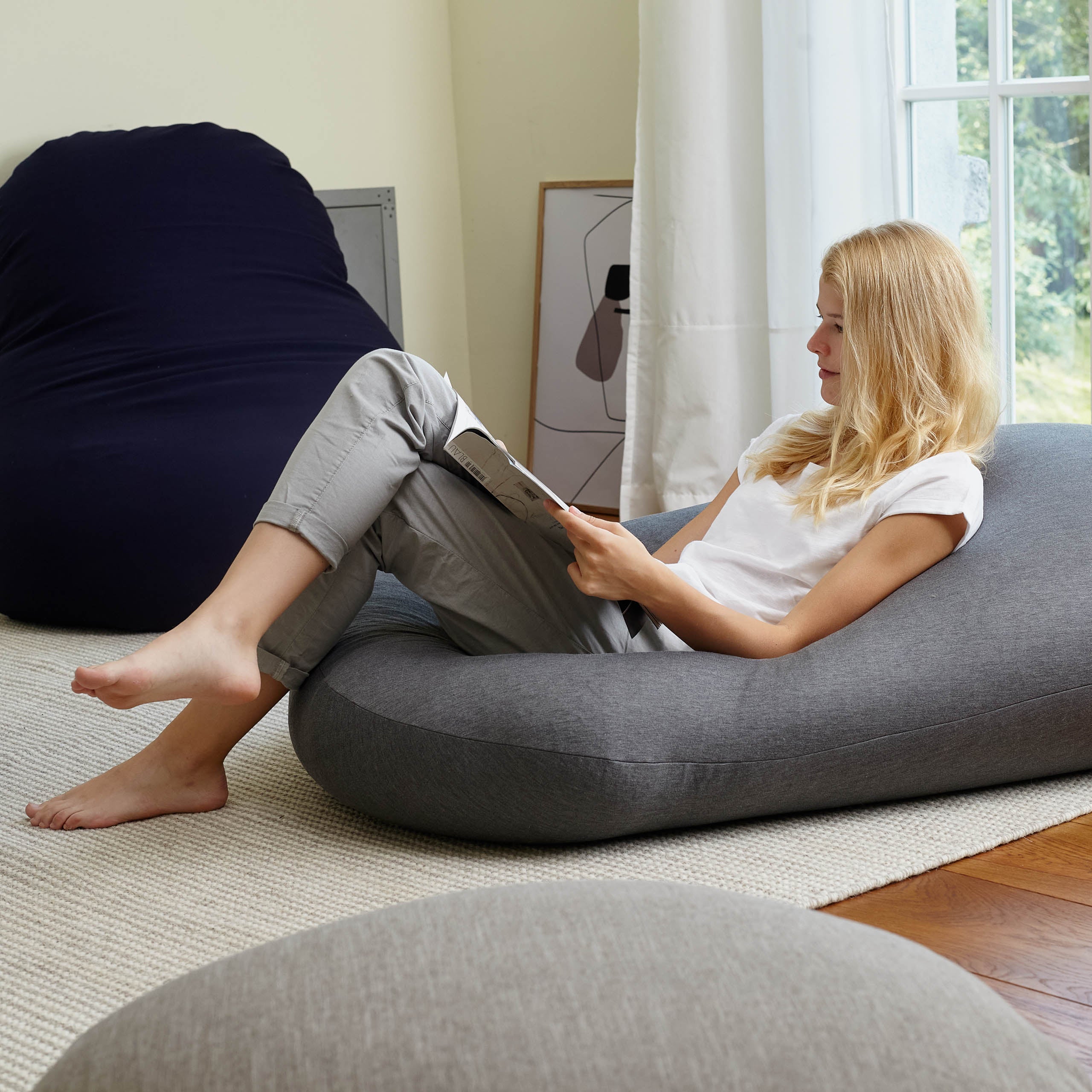 Eine Frau liegt lesend in einem hellen Wohnzimmer auf dem dunkelgrauen Comfort Sitzsack der als Bodenkissen verwendet wird.
