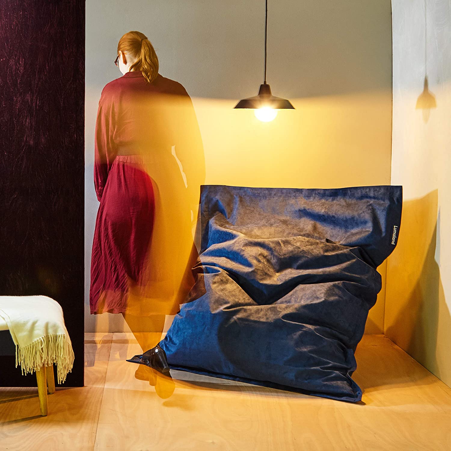 Der XXL Microvelours Sitzsack in Royalblau liegt im Wohnzimmer, eine Frau im roten Kleid geht vorbei.